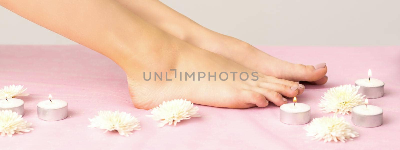 Beautiful white smooth feet of young woman with candles and flowers on pink couch