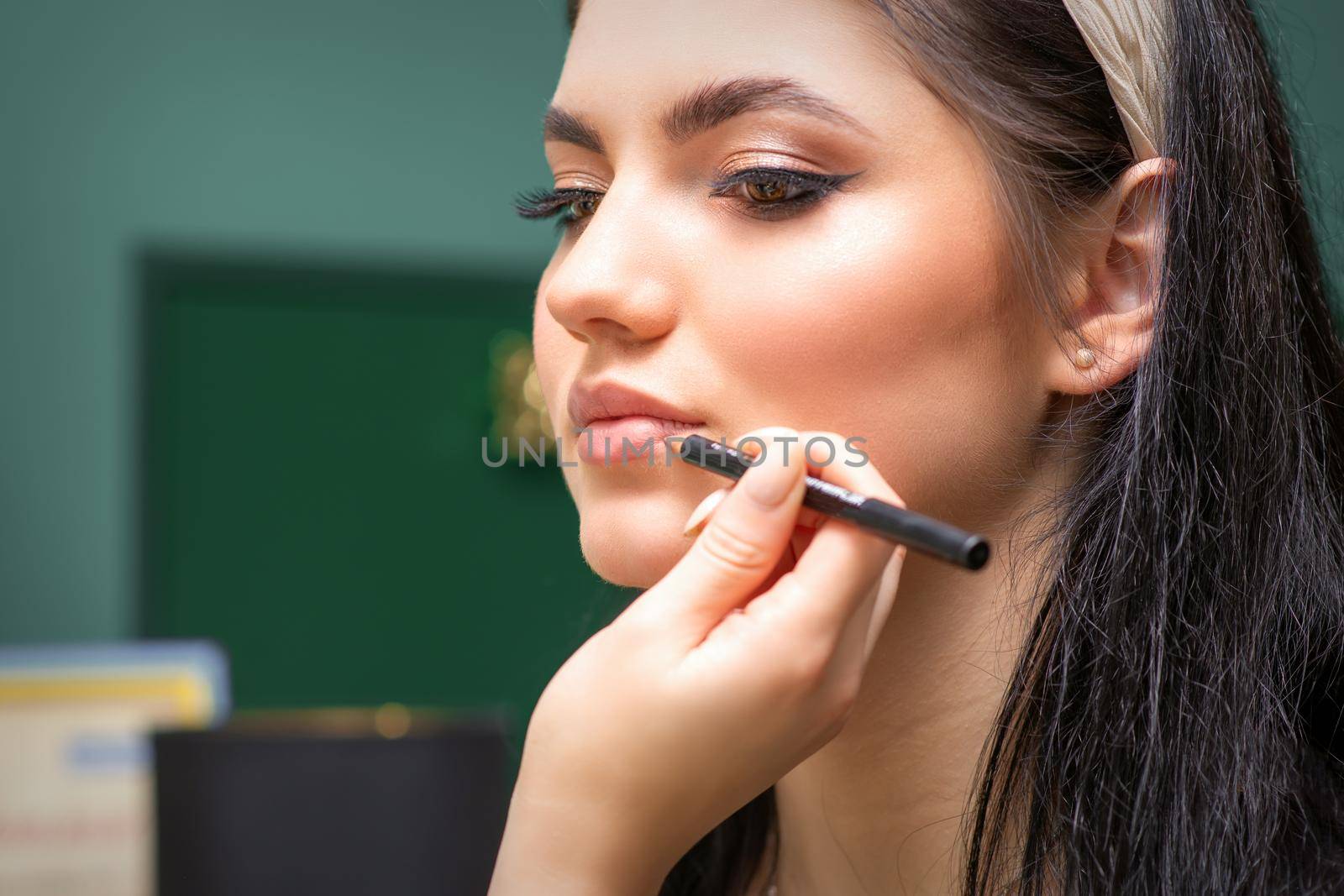 Makeup professional artist or cosmetologist is painting contour lips of a young woman with a pencil close up
