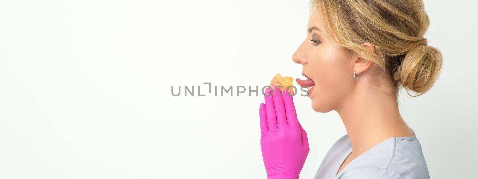 Young beautician licks liquid wax for depilation holding in gloved hands standing on white background. Cosmetologist tastes the sugar paste to taste