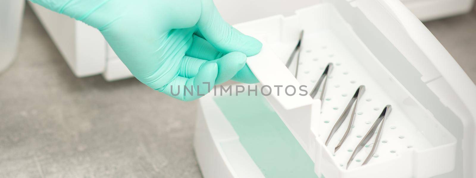 The beautician holds a tray of tweezers preparing them for disinfection with a special liquid and machine