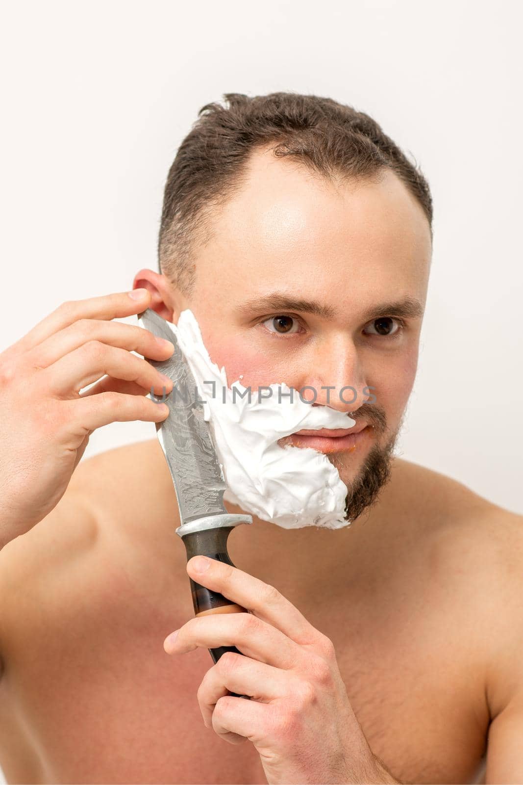 Young caucasian man shaving beard with a big knife on white background