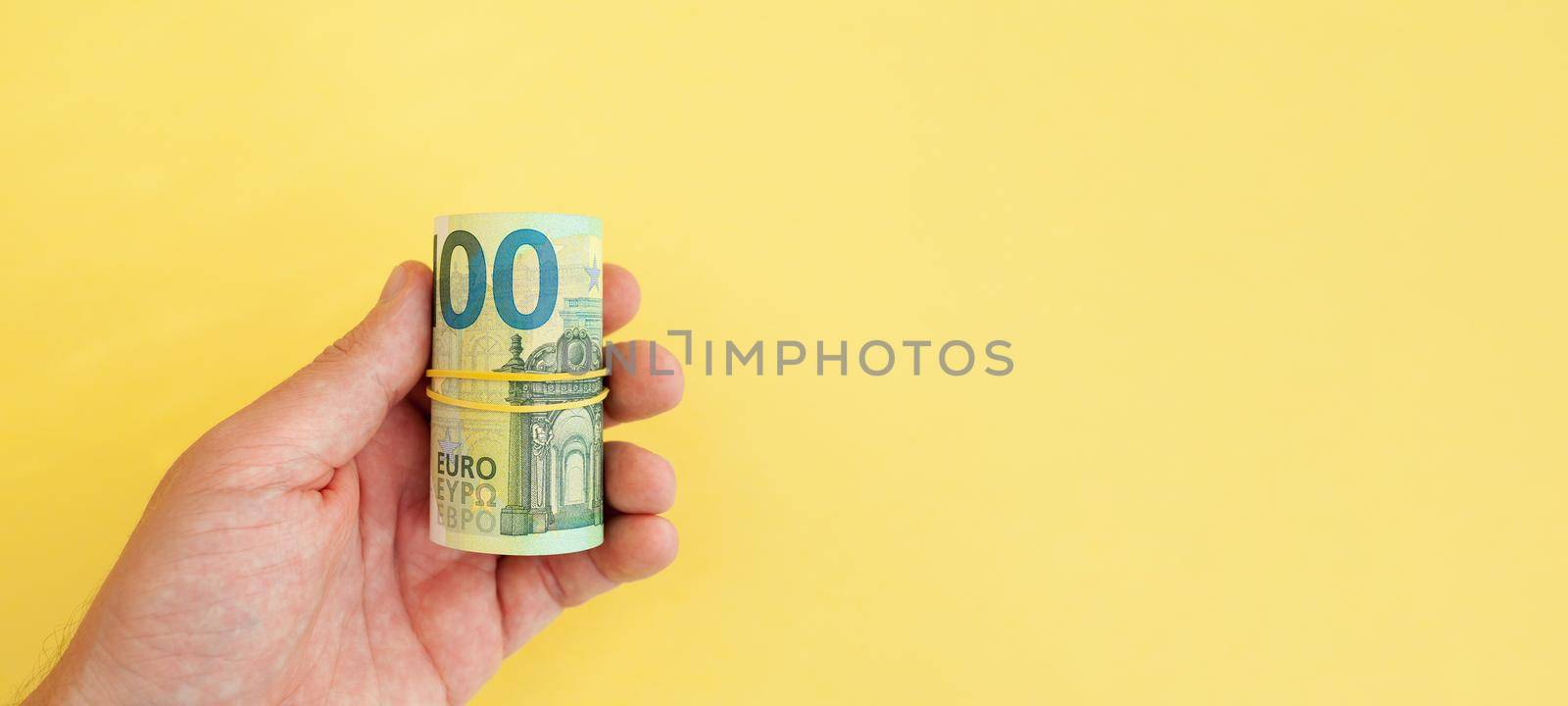 The man holds in his hand money roll of euro banknotes with yellow rubber band on yellow background. cash paper currency, payment, earning and savings, money and finance concept. Empty space for text