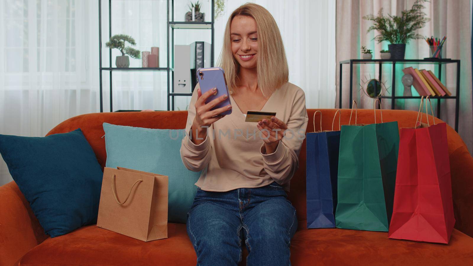 Happy shopaholic consumer girl sitting with shopping bags at modern home apartment making online order, payment with credit plastic bank card. Young woman on couch at home. Discount sale on internet