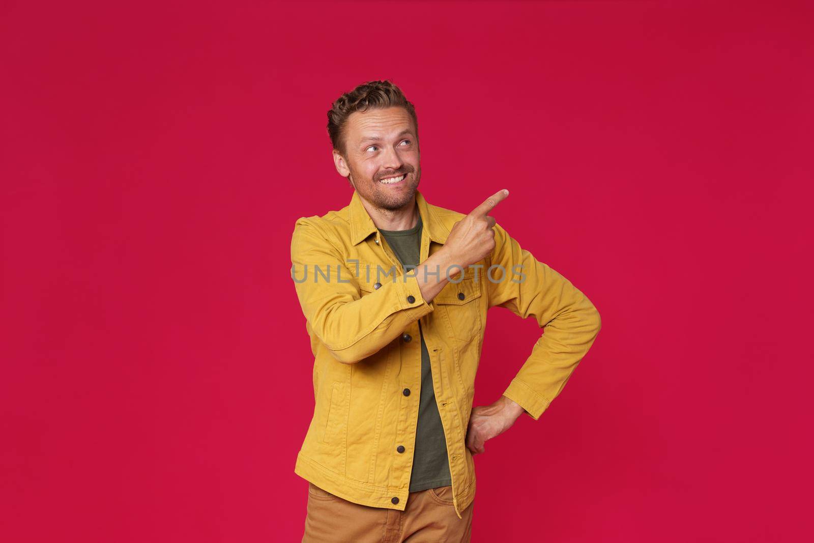 Handsome young man in jeans jacket pointing looking sideways up mock up advertisement isolated on red background. Charming smiling freelancer introduce your business offer.