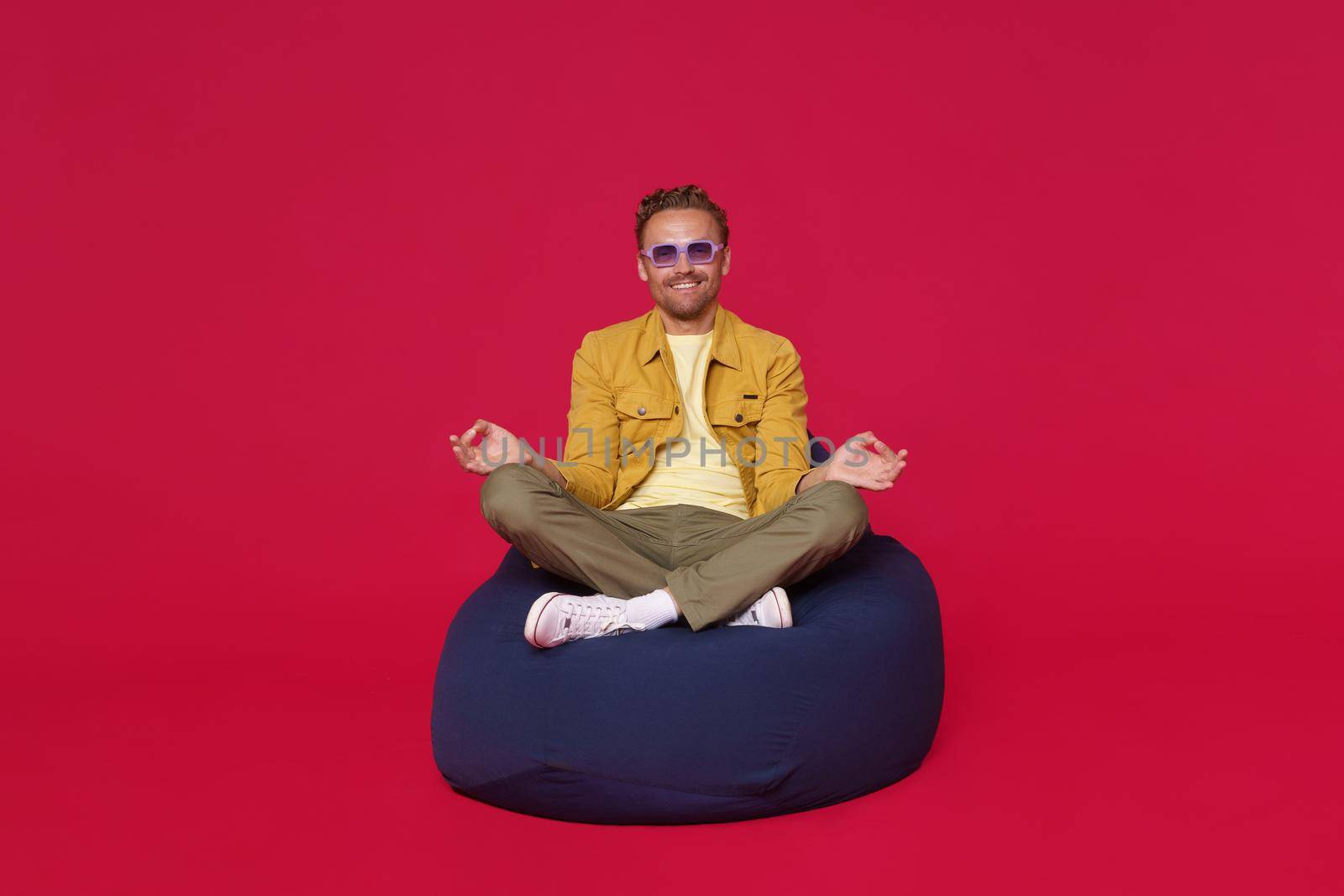 Zen man. Handsome calm focused guy sitting on bag chair in meditating lotus pose wearing blue shades, sunglasses and casual outfit isolated on red background. Happy calm freelancer enjoy free time.