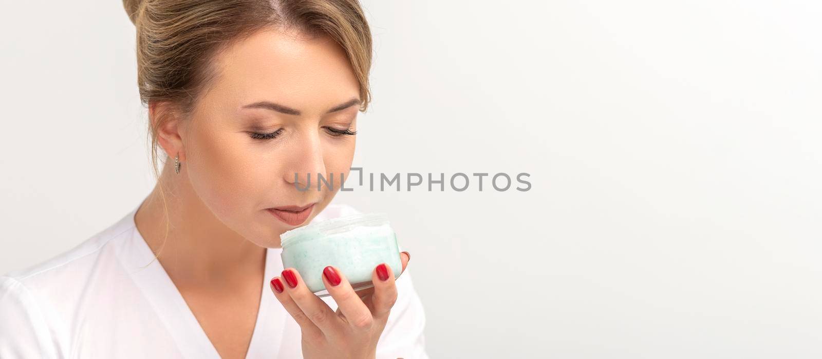 Portrait of young beautician sniffing moisturizing cream holding white jar on white background, copy space