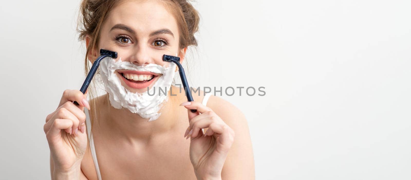 Beautiful young caucasian woman shaving her face by razor on white background. Pretty smiling woman with shaving foam and razor on her face. by okskukuruza