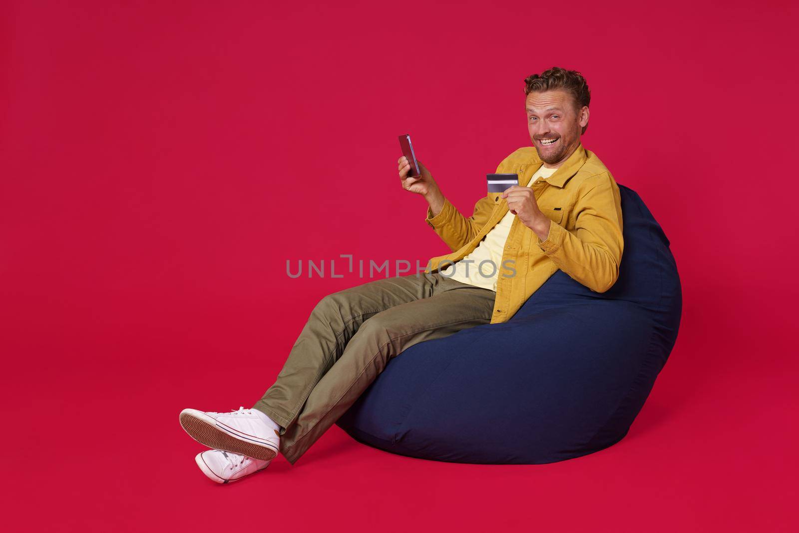 Buying online using debit, credit card handsome young man 30s wearing casual denim clothes sit in bag chair holding digital tablet in hands working isolated on red background. Freelancer man.