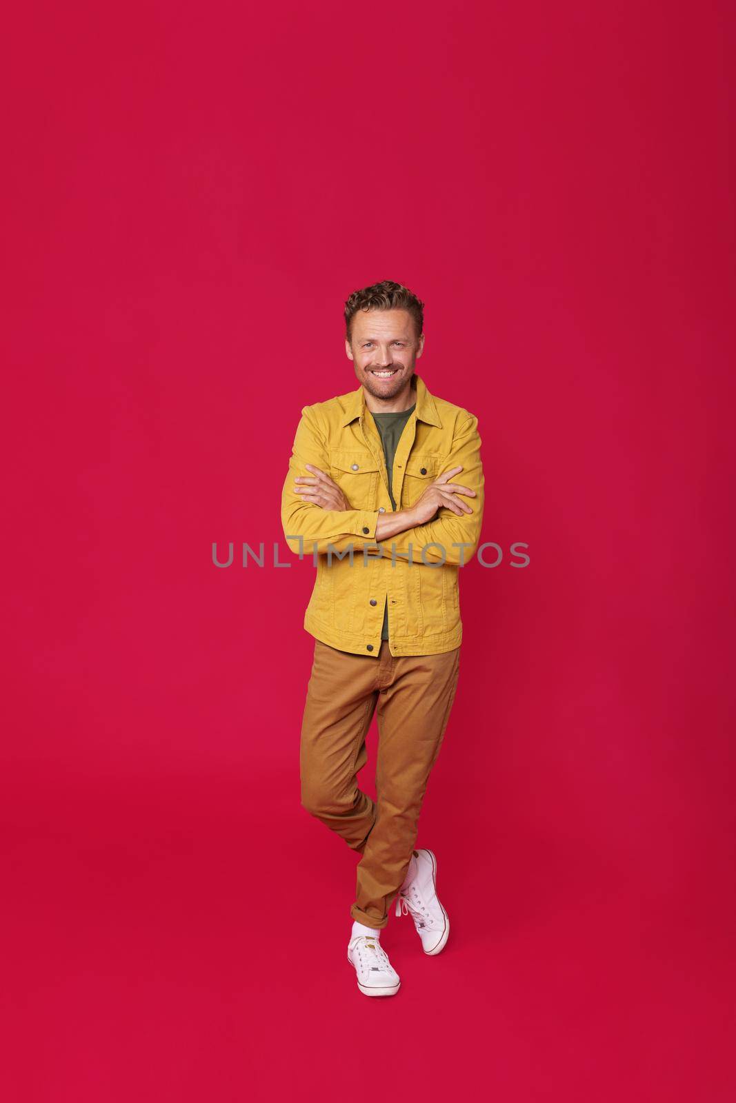 Smiling handsome young man in jeans clothes and white running shoes standing with arms and feet crossed isolated on red background. Happy freelancer man enjoy his success project by LipikStockMedia