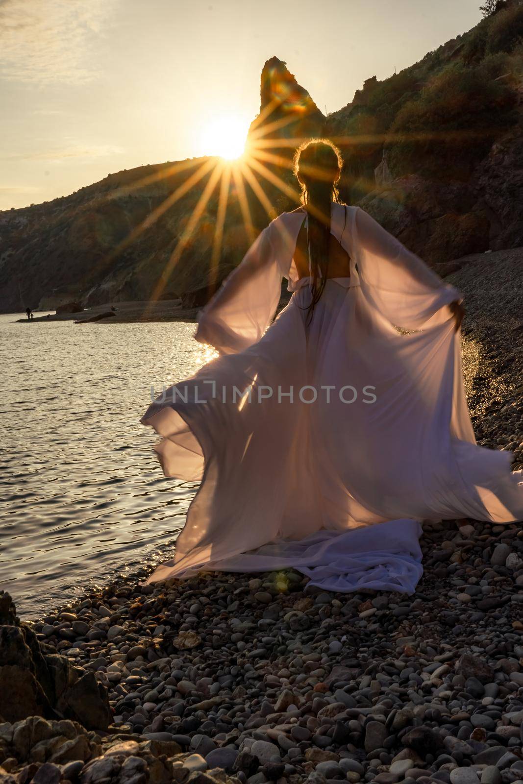 A mysterious female silhouette with long braids stands on the sea beach with mountain views, Sunset rays shine on a woman. Throws up a long white dress, a divine sunset. by Matiunina