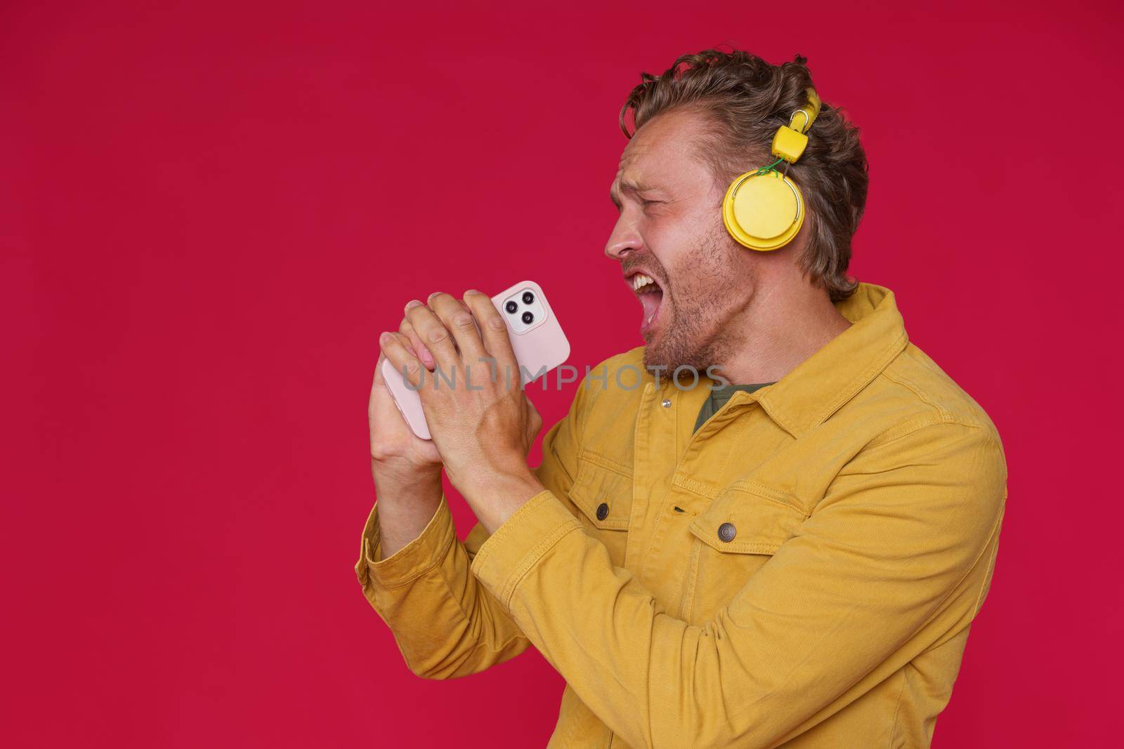 Handsome man 30s singing enjoying his favorite song or track using phone and wireless headphones wearing denim yellow jacket isolated on red background. Joyful man sing while listen music.