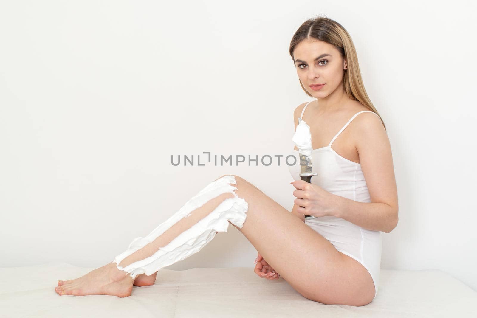 Beautiful caucasian woman shaving her legs with a knife with shaving foam on white background. Depilation and epilation concept