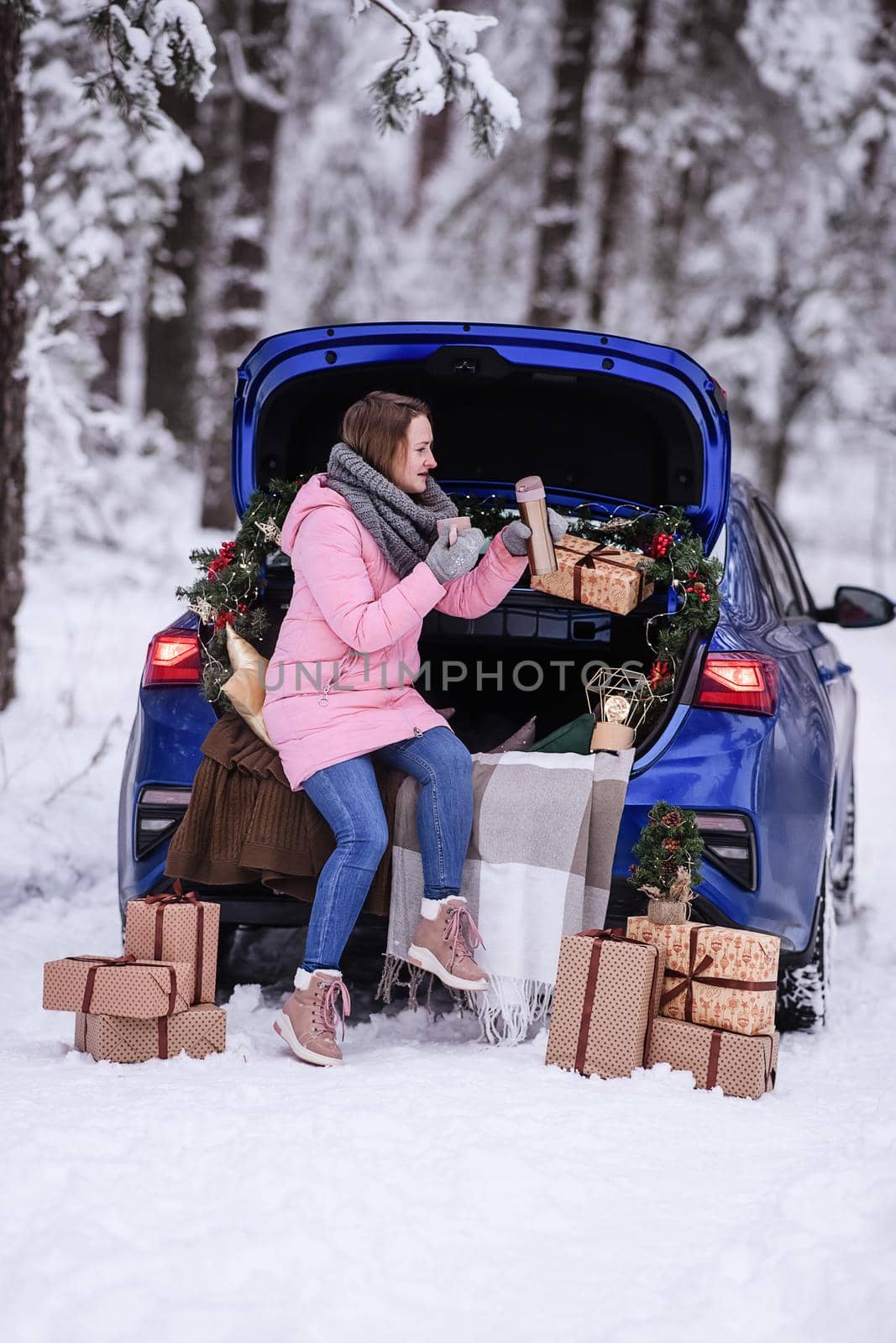 A woman in winter clothes drinks a hot drink sitting in a car decorated in a New Year's way. Traveling by car through the forest. A trip before Christmas.