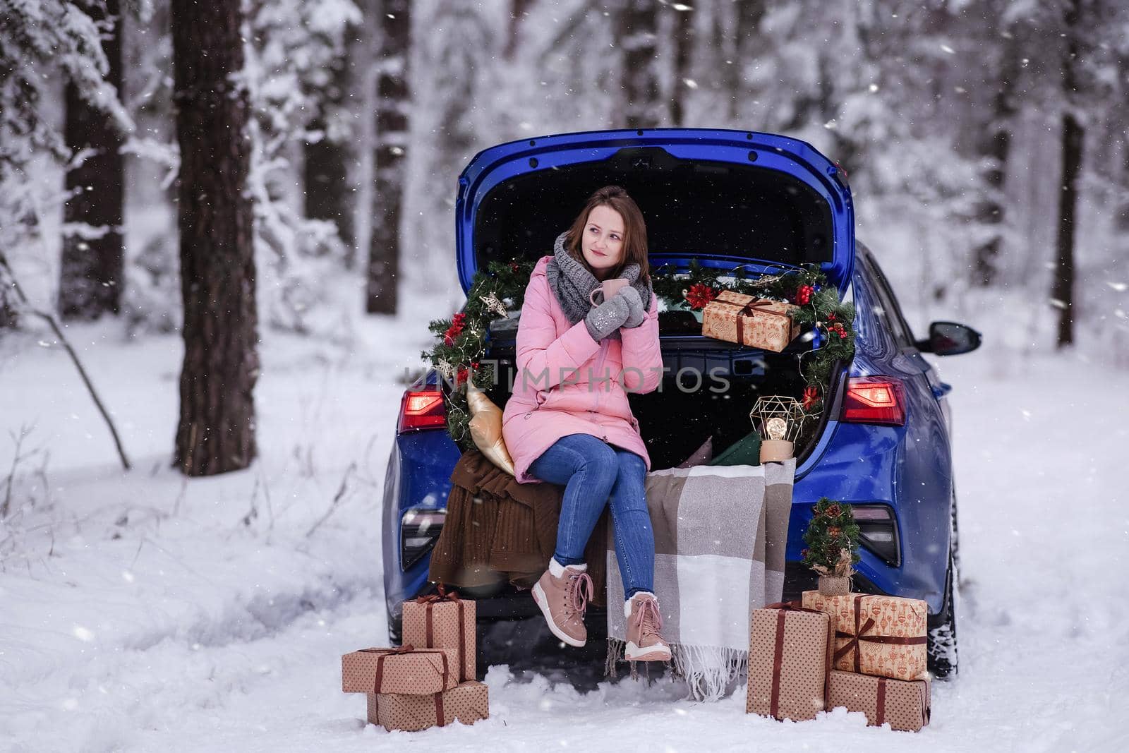 A woman in winter clothes drinks a hot drink sitting in a car decorated in a New Year's way. Traveling by car through the forest. A trip before Christmas.