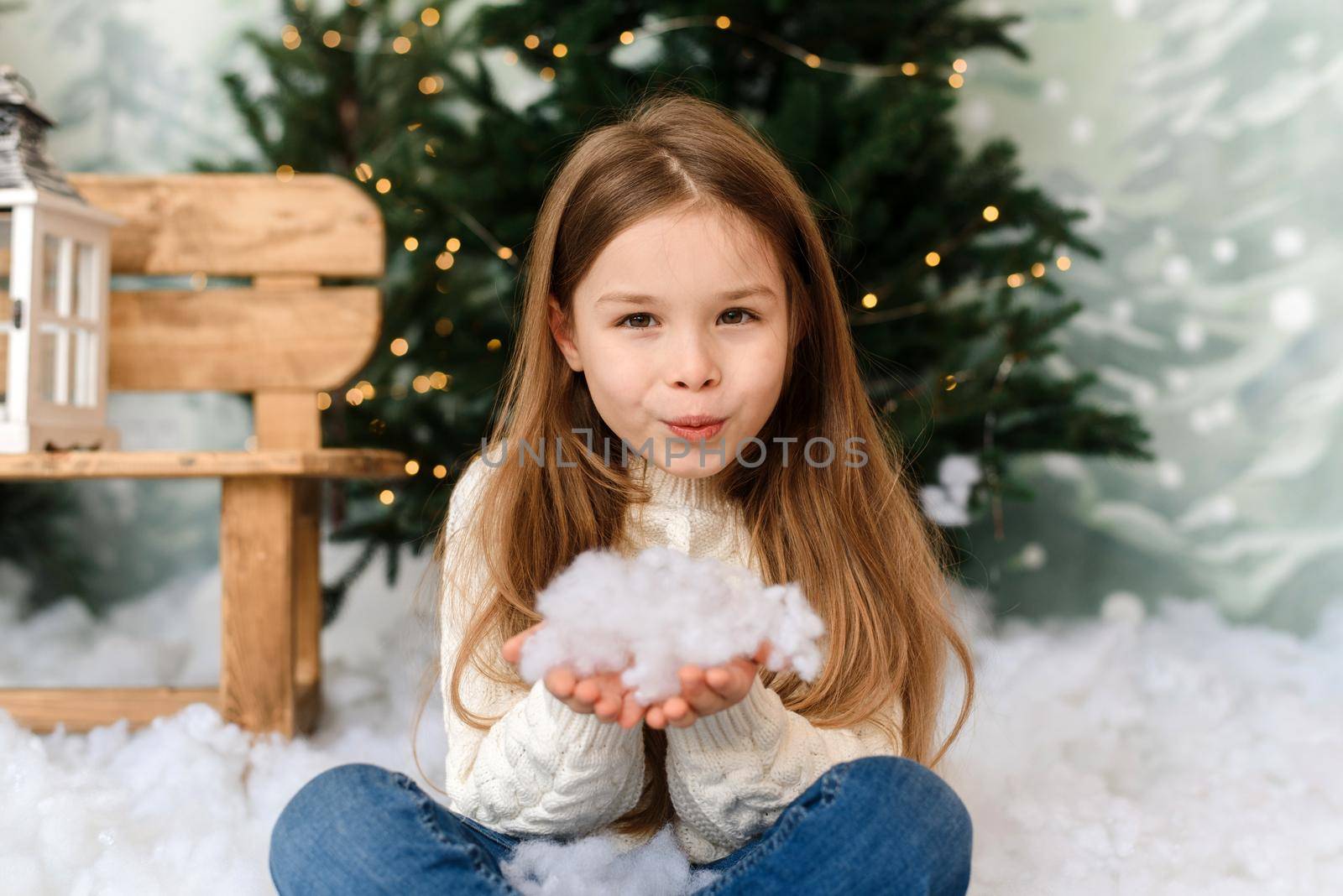An adorable little girl in the interior of a Christmas studio blows artificial snow into the camera and rejoices at Christmas Eve