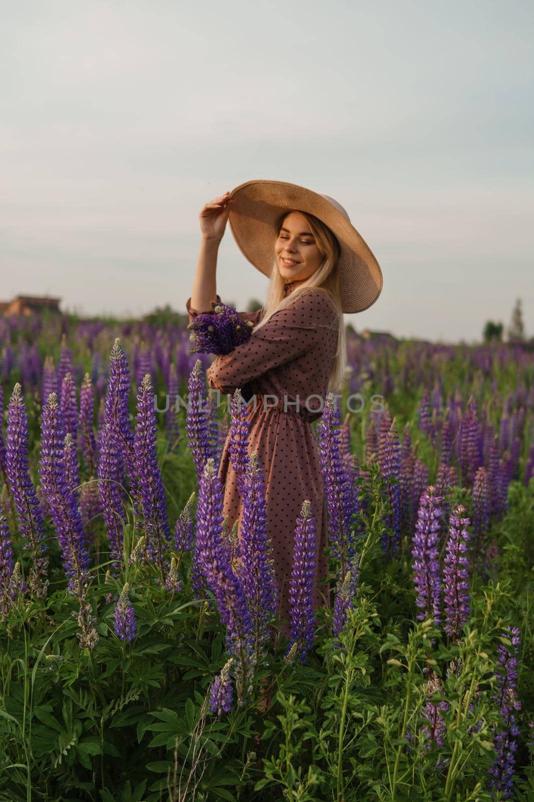 A beautiful woman in a straw hat walks in a field with purple flowers. A walk in nature in the lupin field.