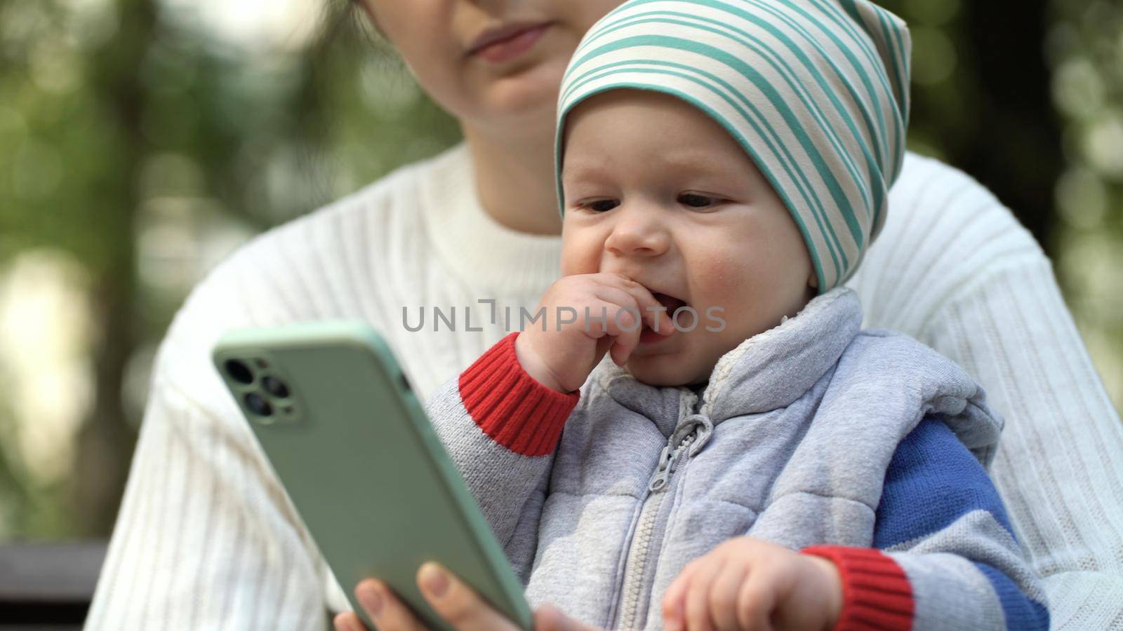 A one-year-old boy sits in his mother's arms in the park and watches a cartoon on the phone in the park close-up. Happy child waving hands while watching cartoons on smartphone by Petrokill