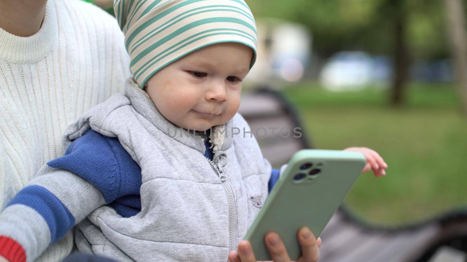 Portrait of a joyful baby boy watching a cartoon on the phone in the open air and waving his hands with joy.