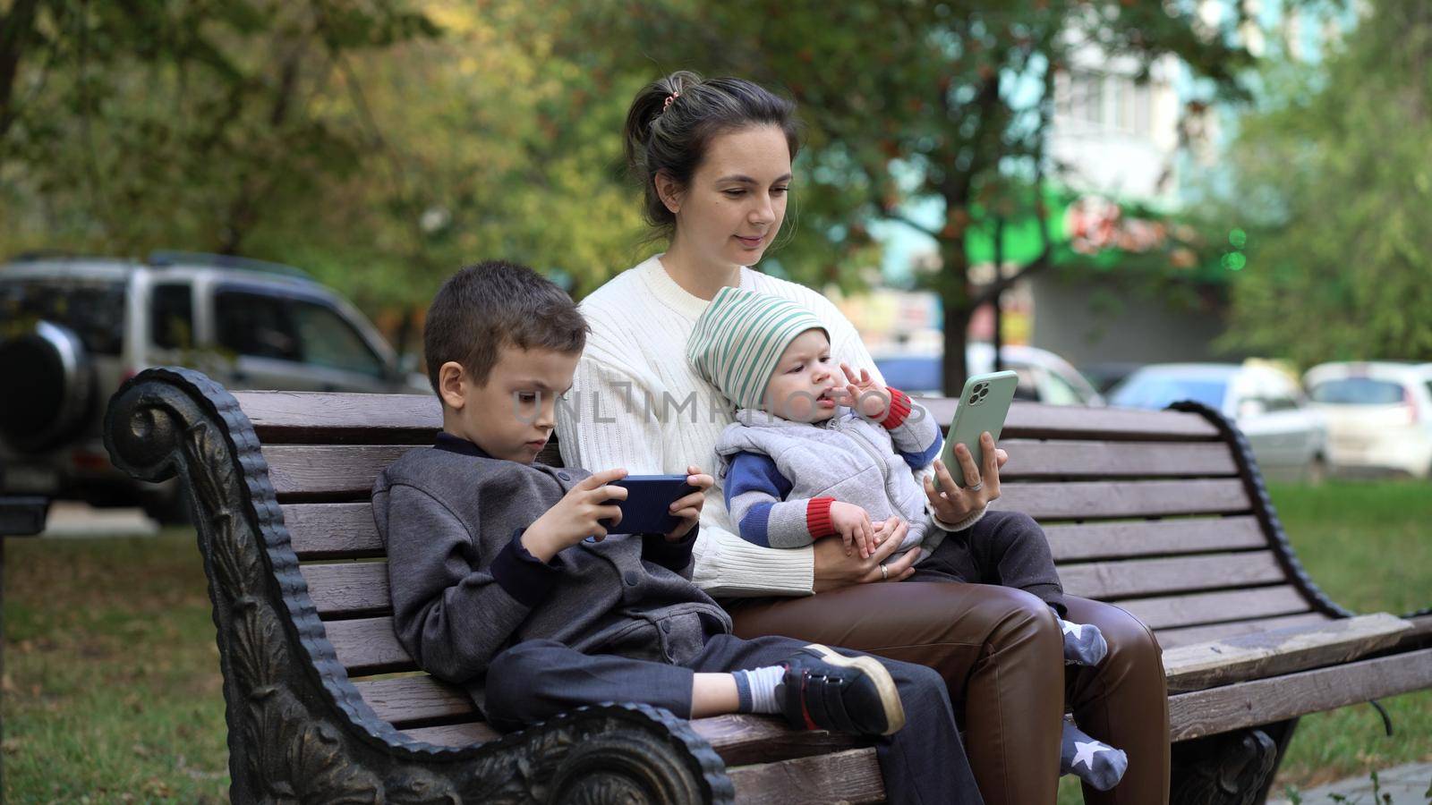 Mom with two children are sitting on a bench in the park and are passionate about playing on the phone and watching cartoons.