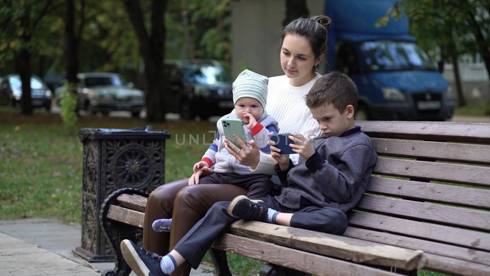 Modern family relaxing with gadgets outdoors on a bench outside. Mom and two sons in the park by Petrokill