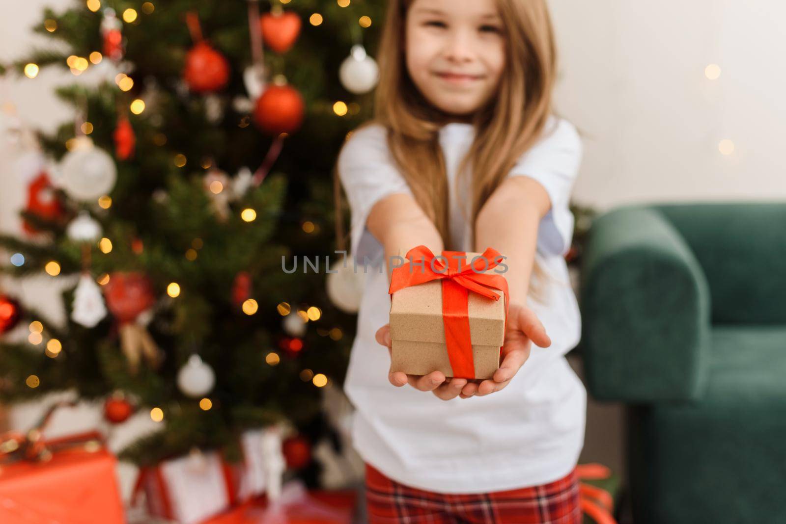 Cute girl in pajamas gives a gift to the camera on the background of the Christmas tree.