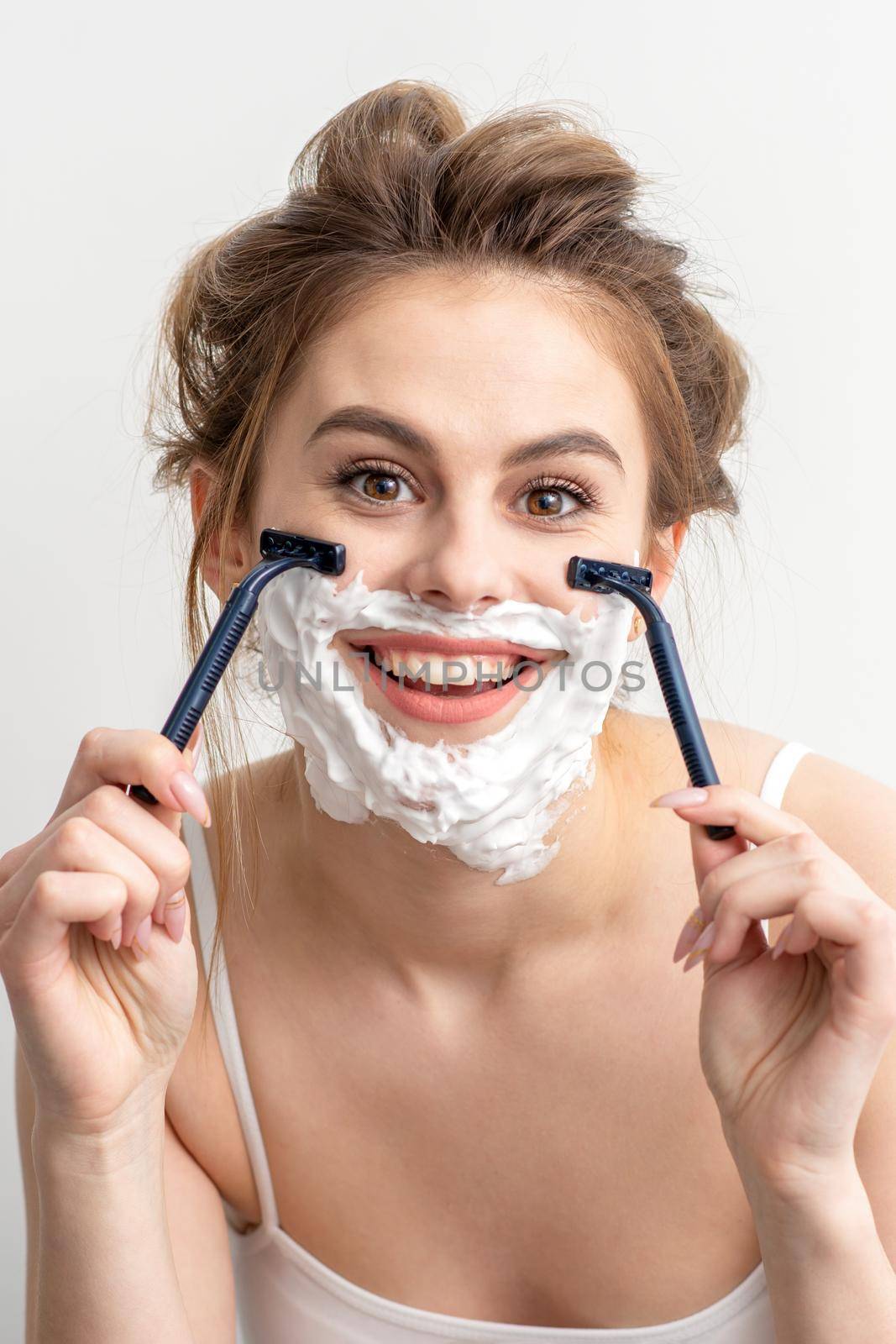 Beautiful young caucasian woman shaving her face by razor on white background. Pretty smiling woman with shaving foam and razor on her face. by okskukuruza