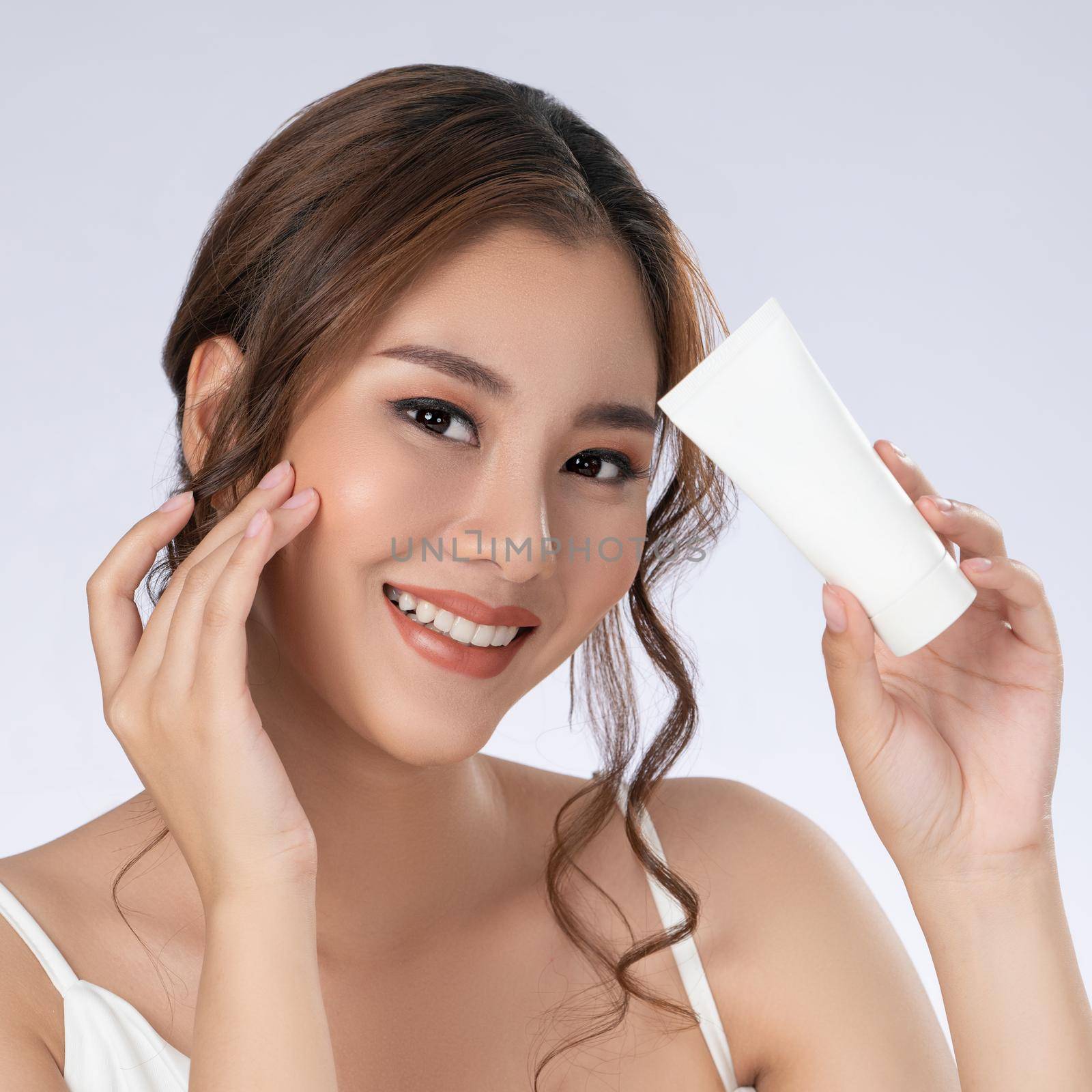 Gorgeous woman with makeup smiling holding mockup product for advertising text place, light grey background. Advertising concept for skin healthcare and beauty care products.