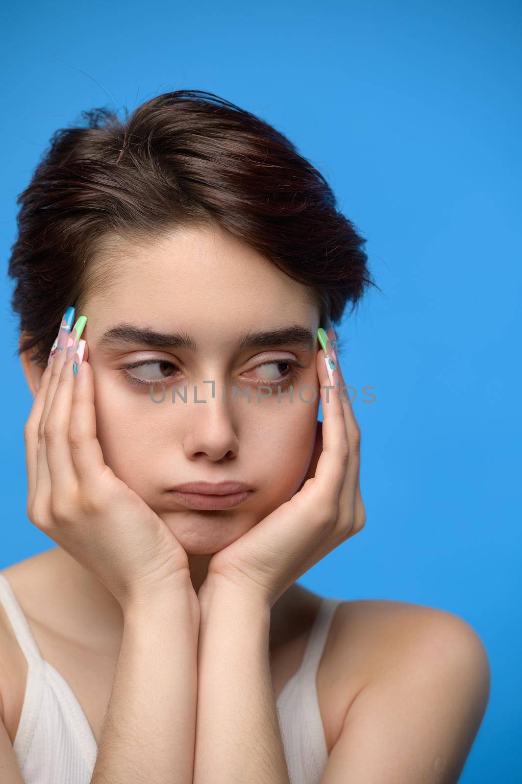 Bored and sulking sensual young teenage girl with shork hair and long extravagant nailart looking sideways, at blue background