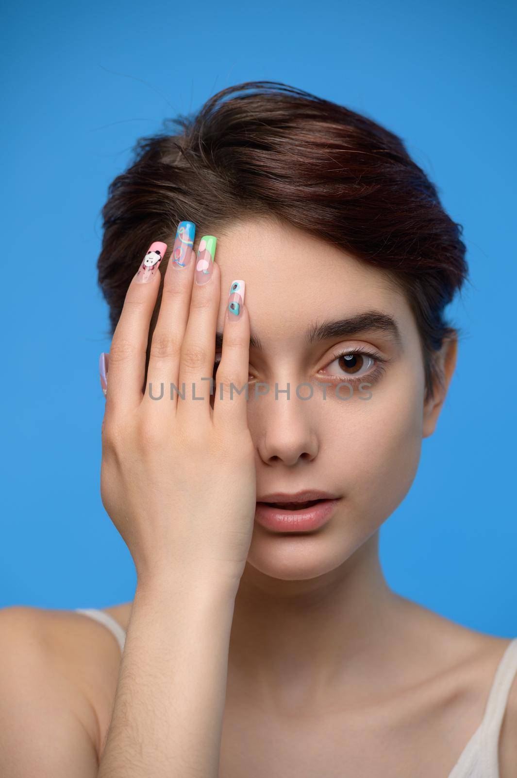 A skinny beautiful young brunette with her palm covering the right side of her face, at blue background