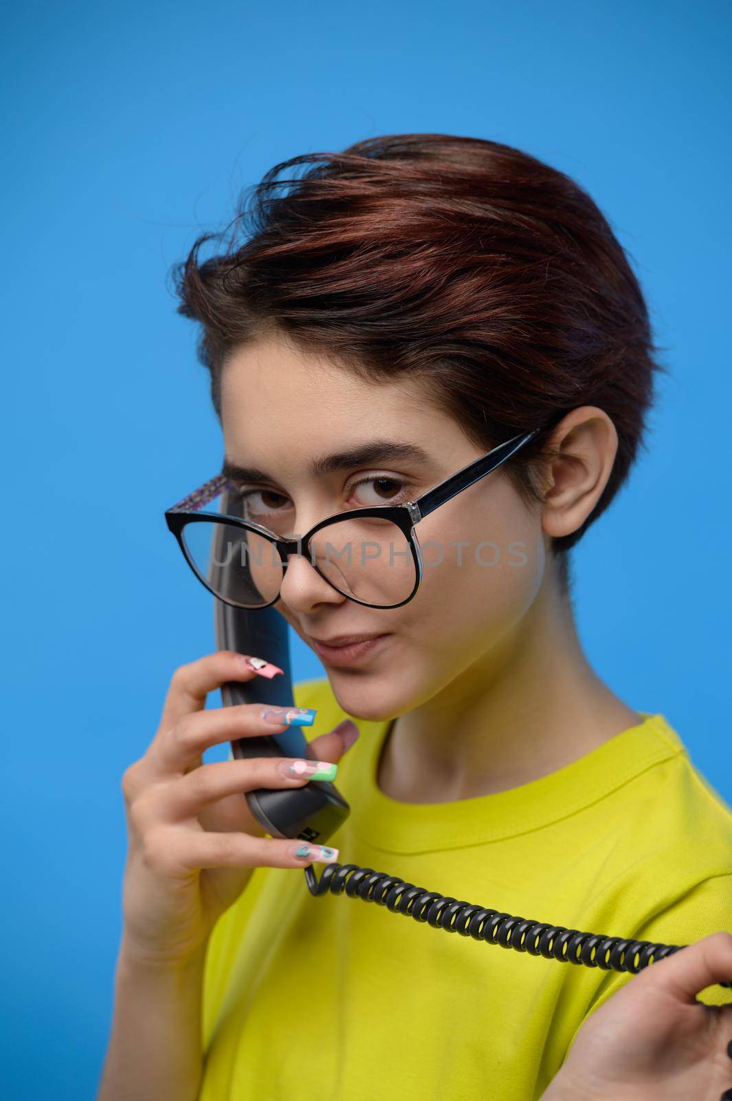 Smiling young pretty brunette guirl with extravagant nail art having conversation using old fashioned phone, studio shot at blue background