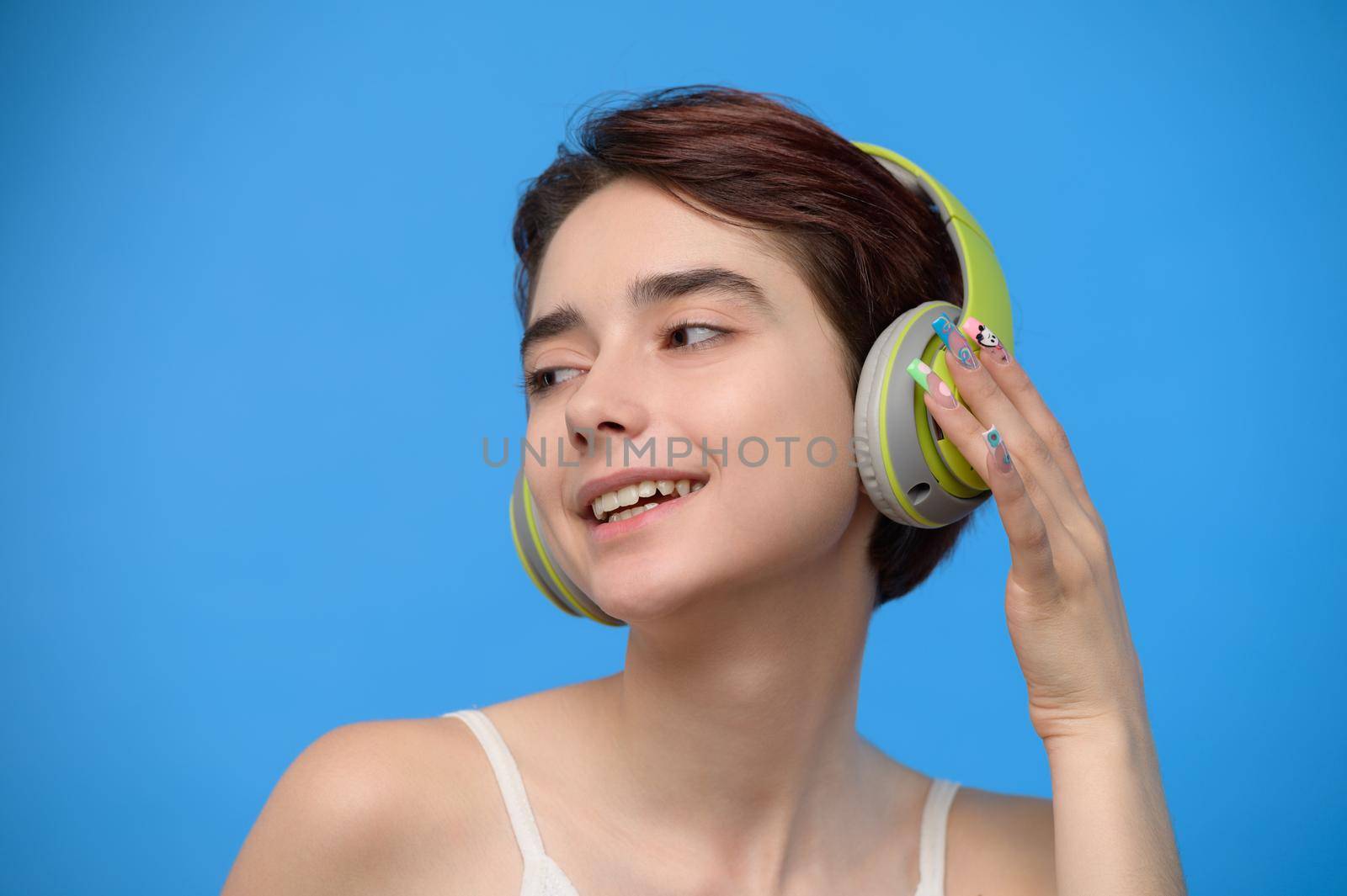 Portrait of cute young bare shoulders brunette with extravagant nail art listening to music using wireless headphones, studio shot