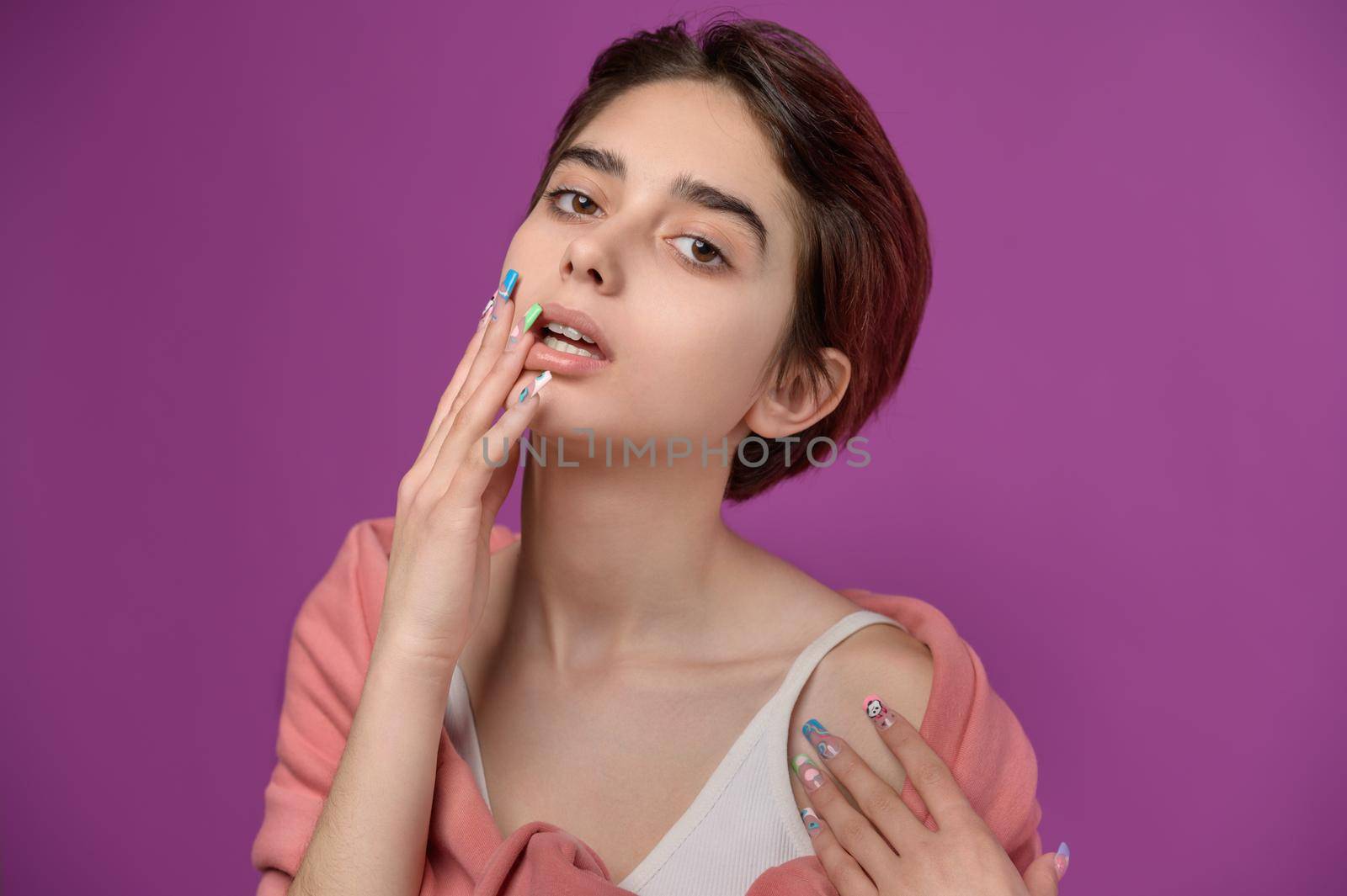 Sensual studio portrait of a young delicate skinny brunette girl, showing an exravagant nail art on her right arm. On a purple background.