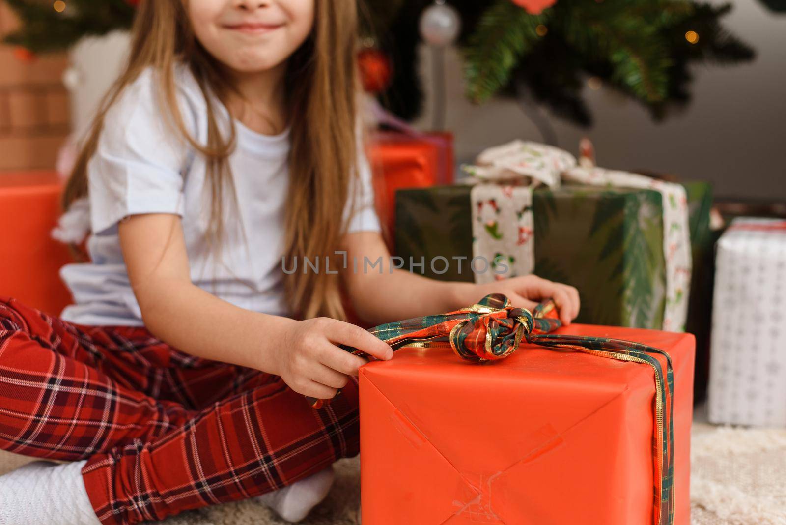 Merry Christmas. Cute little girl opens a present near the Christmas tree. The girl enjoys the gift. by etonastenka