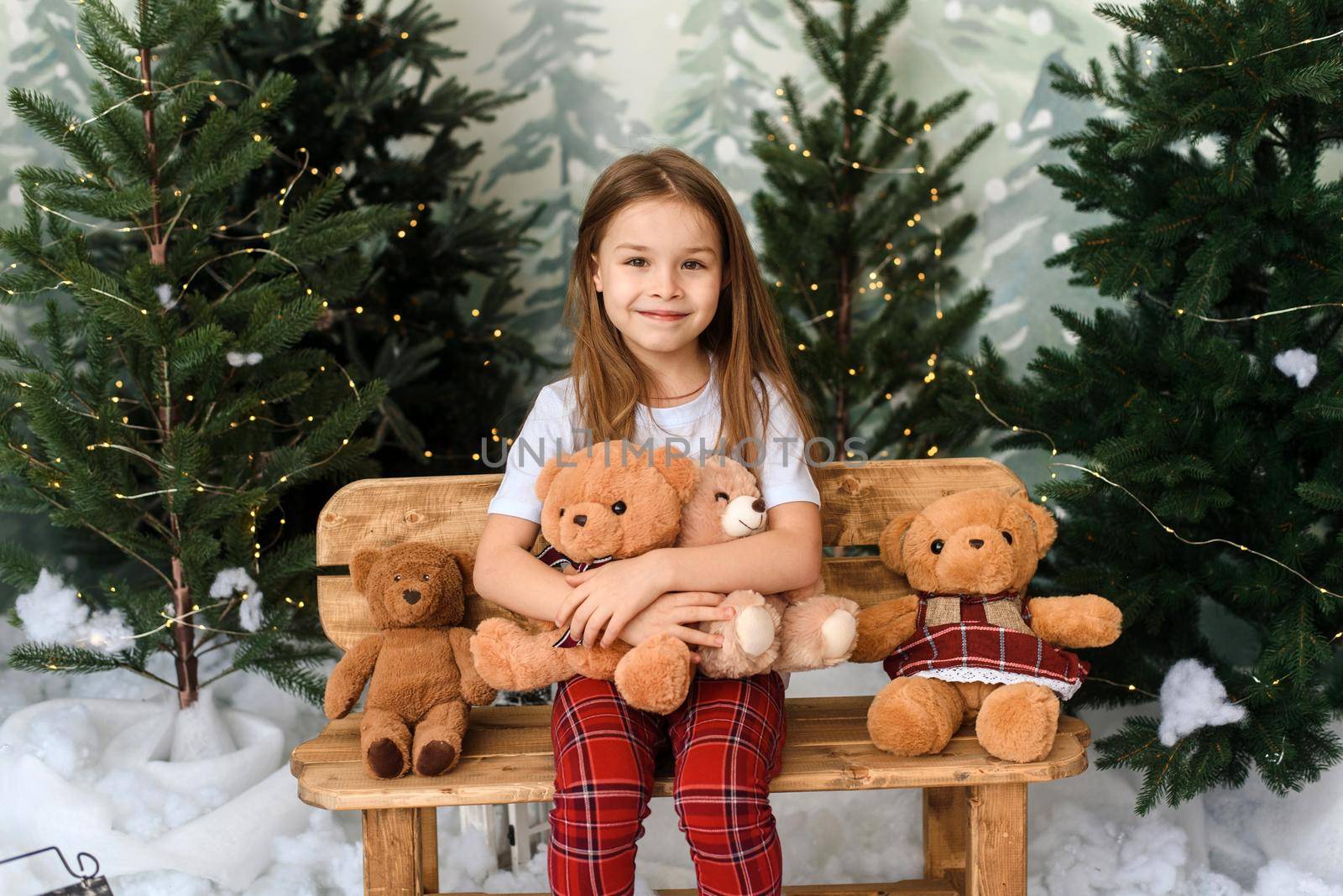 A cute little girl sits on a bench among the Christmas trees and rejoices at the gifts. Child girl with teddy bears in a Christmas interior by etonastenka