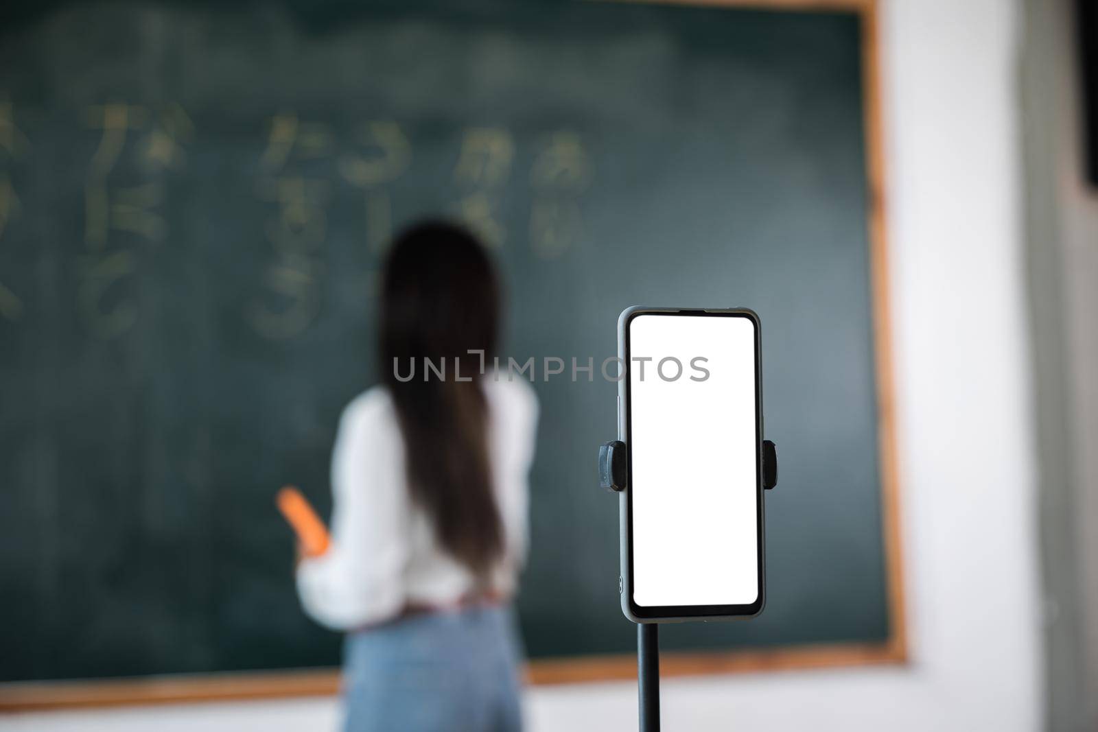 Asian teacher teach on board while have blackboard lecture online through smartphone in classroom by Sorapop