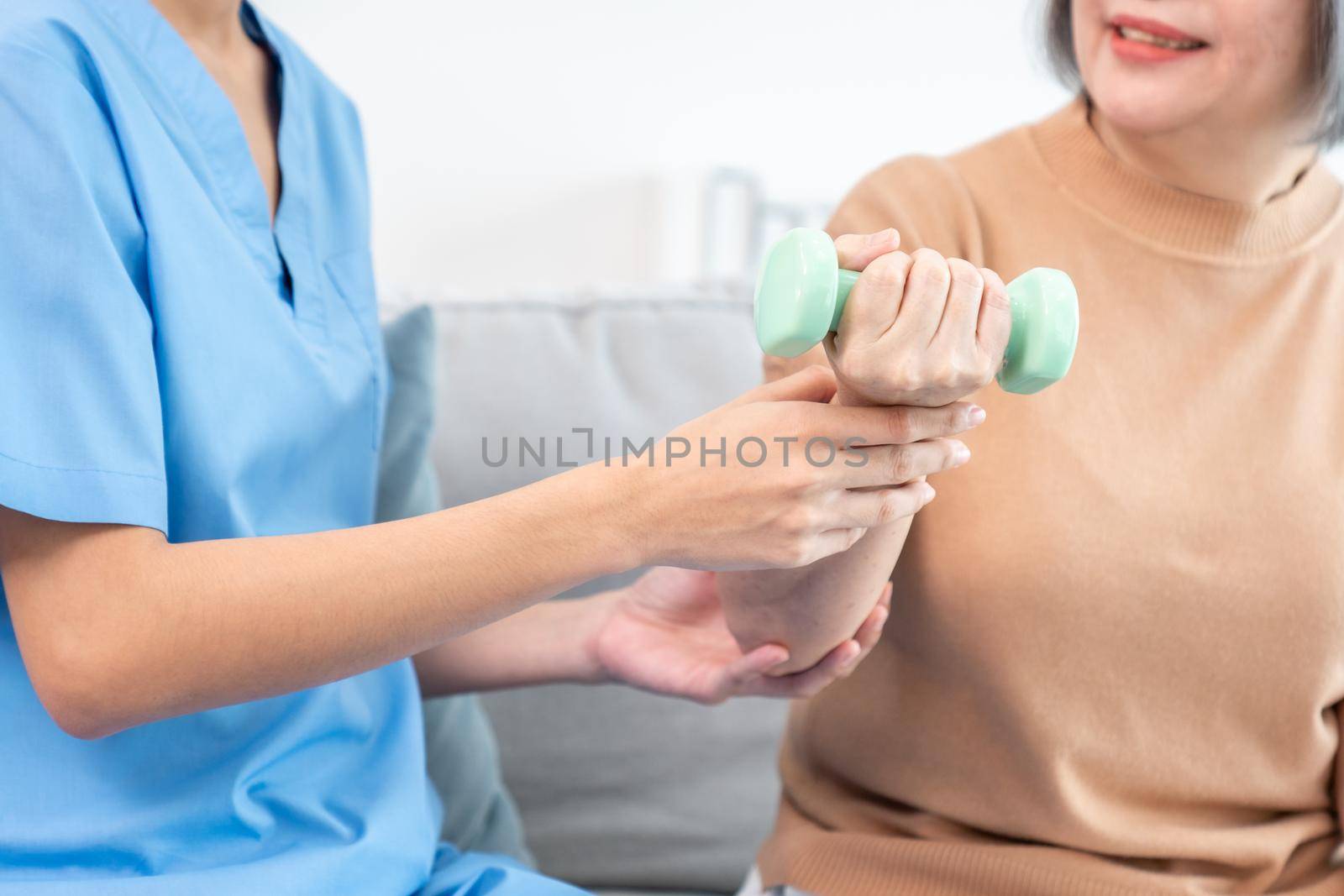 Caregiver helping contented senior woman exercise with dumbbell at home. Rehab for elder, senior medical care.