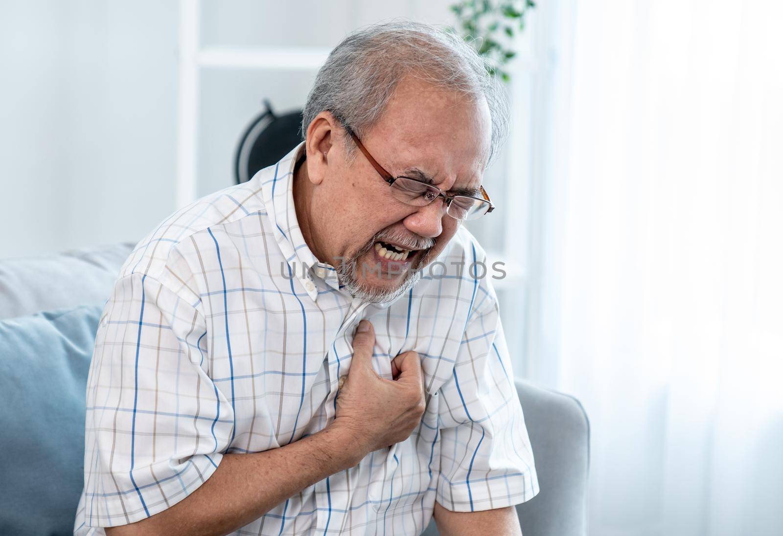 An agonizing senior man suffering from chest pain or heart attack alone in his living room. Serious health problem and feeling unwell concept for seniors.