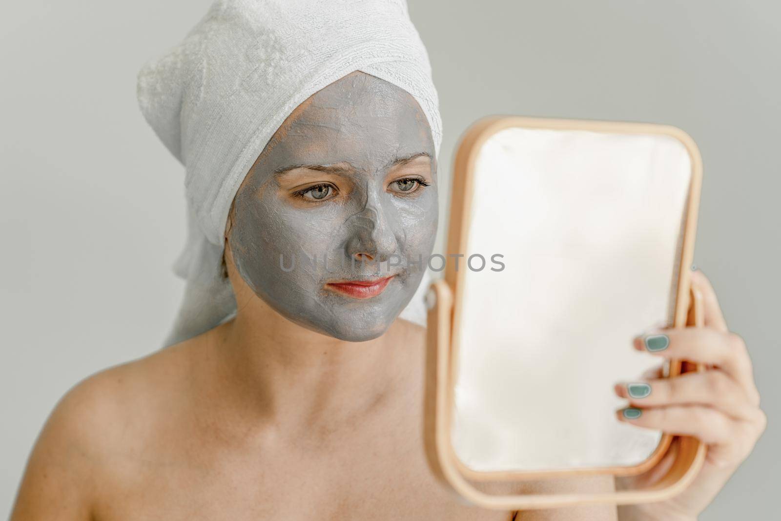 Young woman with gray clay cosmetic mask on her face looks in mirror, her hair and body wrapped in towel. Close-up