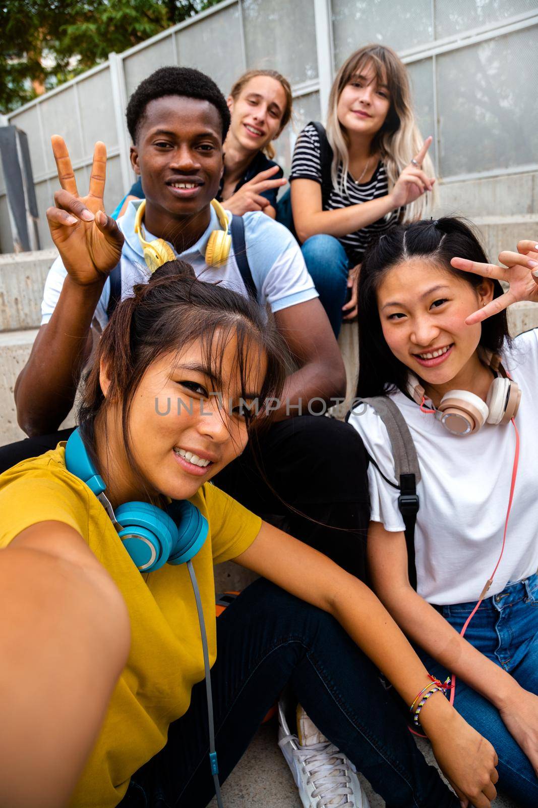 Happy smiling teen multiracial group of students looking at camera take selfie sitting on steps outside. Vertical image. by Hoverstock