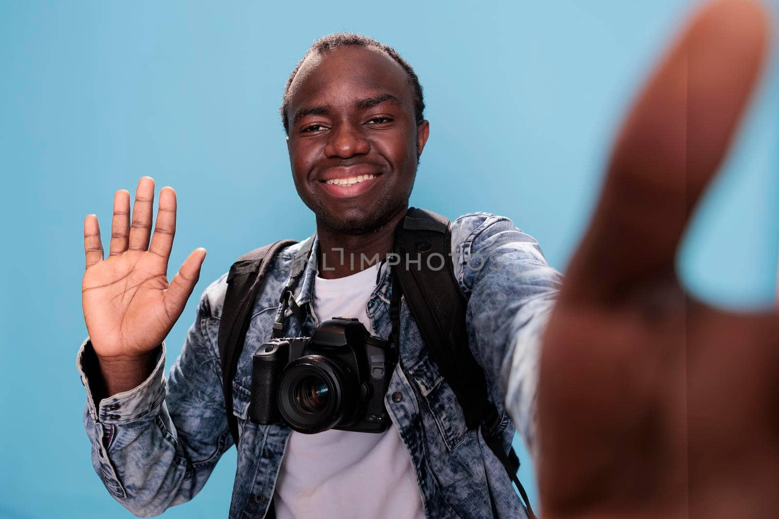 Smiling young man waving at camera while taking selfie photo on blue background. Happy photographer with DSLR device and trip backpack taking picture of himself. Studio shot