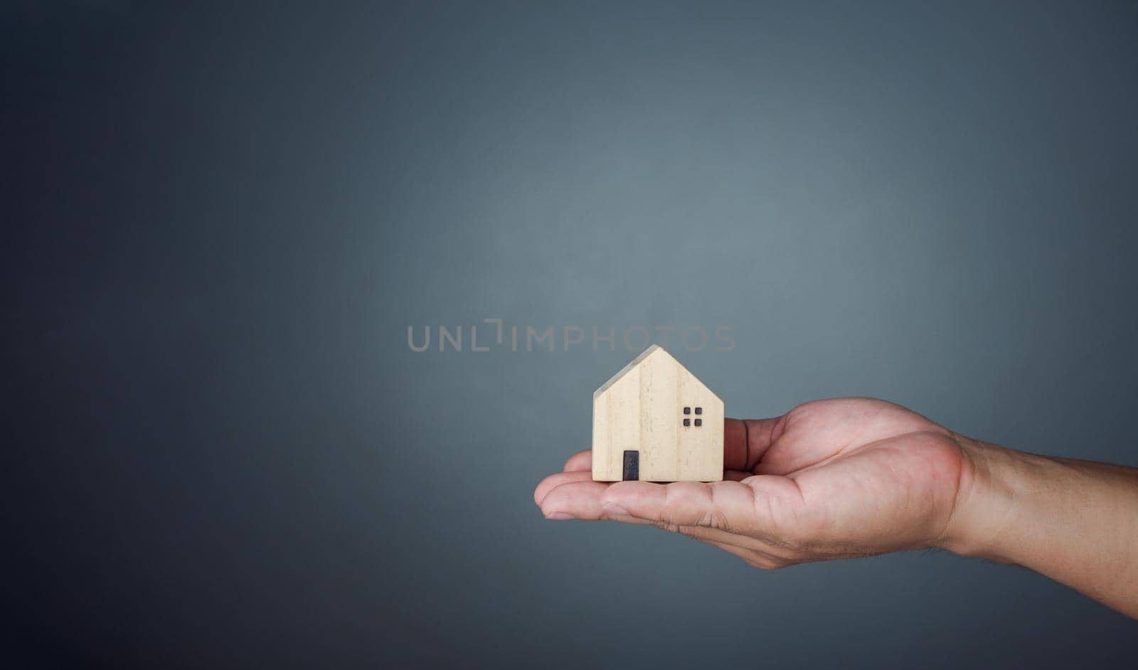 Concept of selling a house. A hand is holding a model house on a gray background. Real estate agent offer house, property insurance and security, affordable housing concepts, home insurance broker agent, salesman person. by Unimages2527