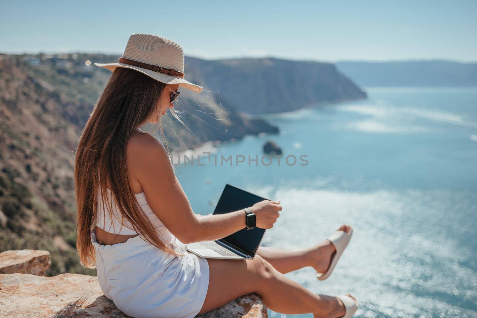 Successful business woman in yellow hat working on laptop by the sea. Pretty lady typing on computer at summer day outdoors. Freelance, travel and holidays concept.