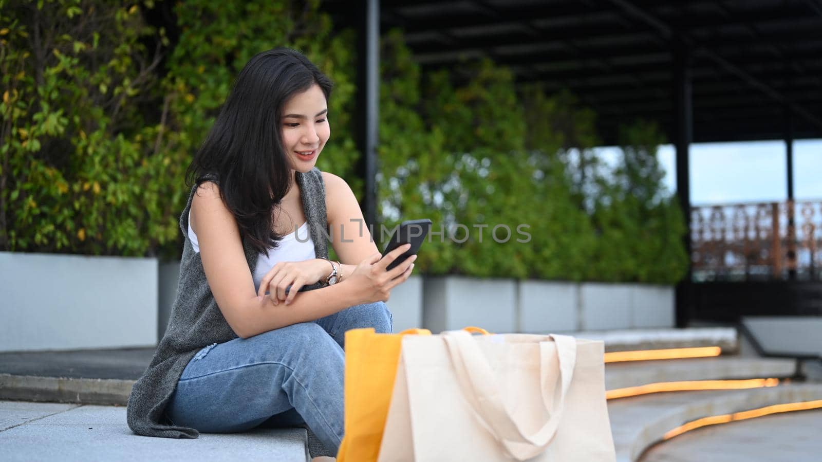 Beautiful asian woman with shopping bags using mobile phone while sitting on stairs in front of the building by prathanchorruangsak