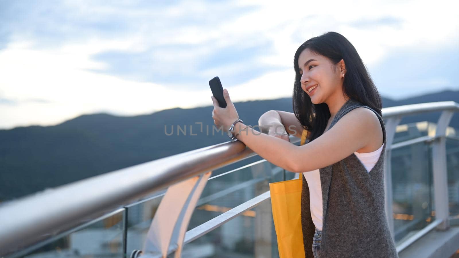 Smiling young woman typing text message on mobile phone while standing on roof terrace outdoors with cityscape on background.