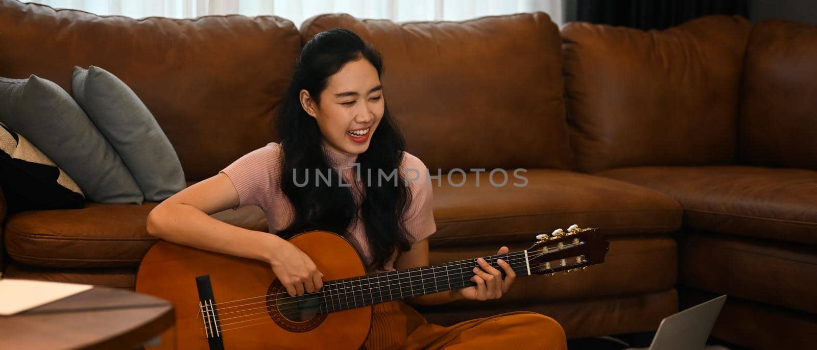 Happy young woman learning play guitar, watching online lessons on laptop. Hobbies and learning concept.