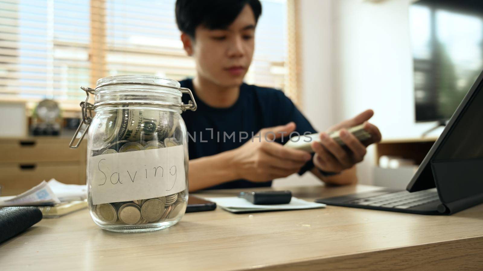 Cropped view of glass jar with coins on wooden table and man using laptop in background. Saving money and investing concept.