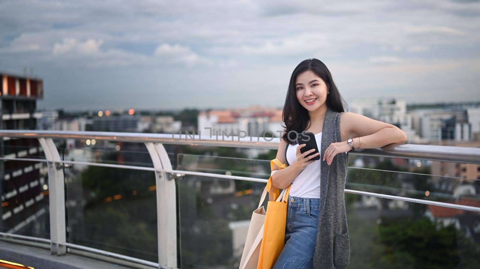 Attractive asian woman holding mobile phone standing on roof terrace outdoors with cityscape on background by prathanchorruangsak