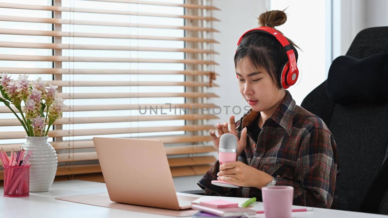 Asian woman using microphone and laptop computer recording podcast at small broadcast studio.