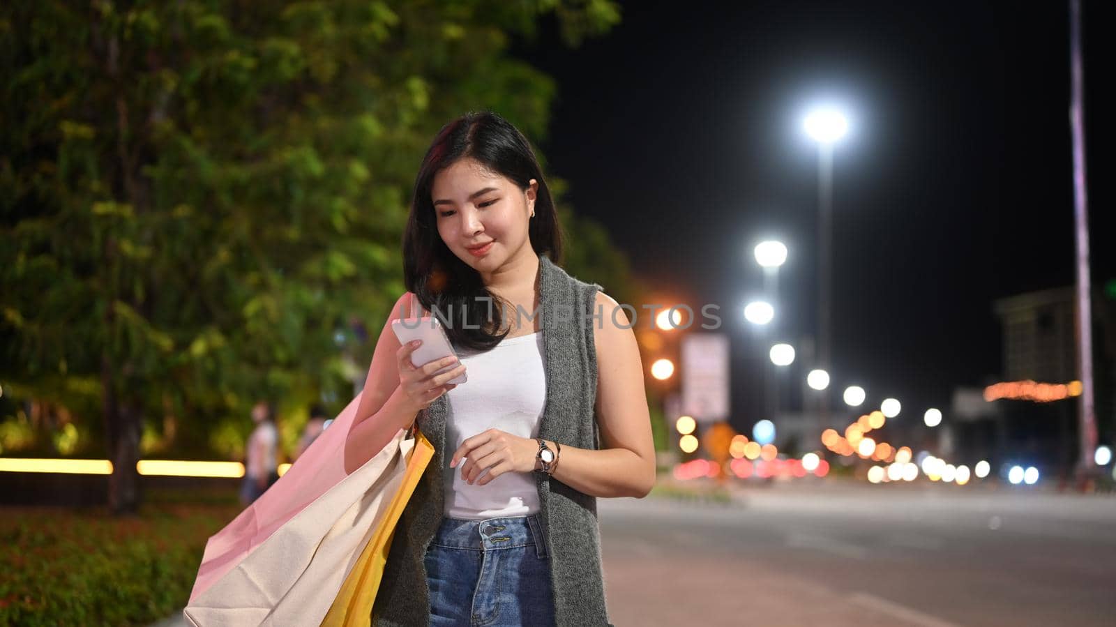Stylish woman typing text message on her mobile phone while walking through night city street with blurred lights background.