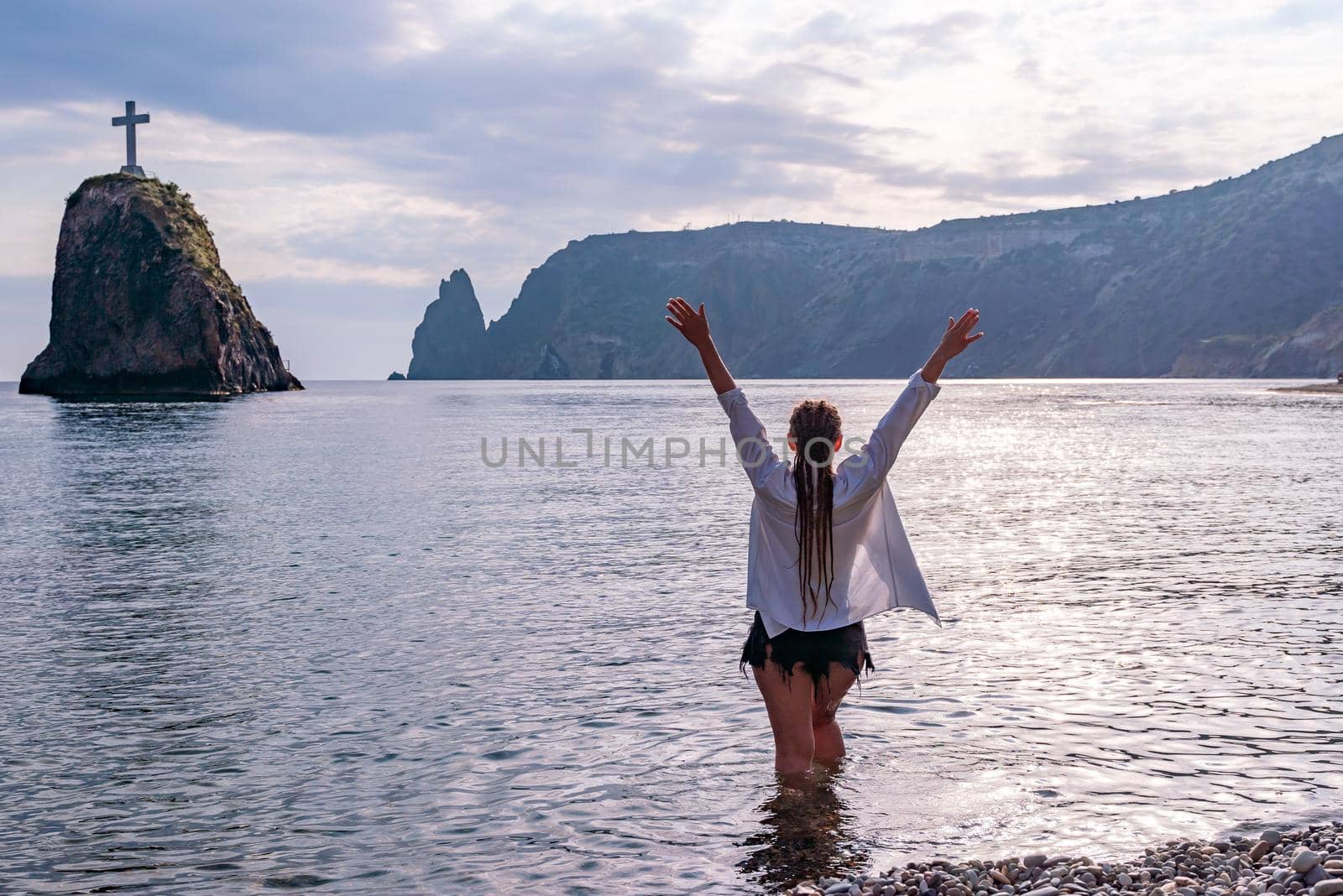 The girl stands on the shore and looks at the sea. Her hands are raised up. She wears a white shirt and her hair is in a braid. by Matiunina